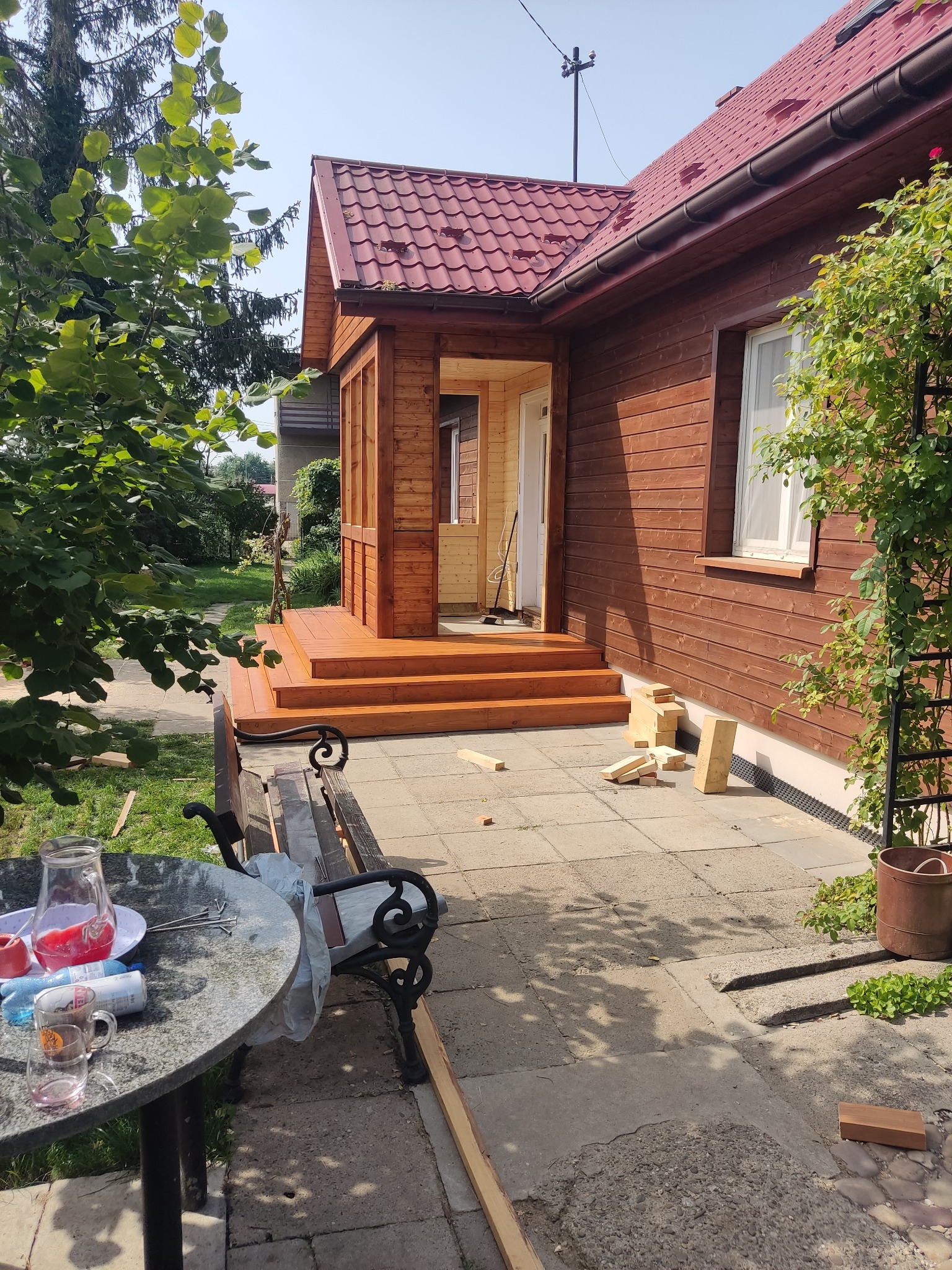 Nowo wybudowany, drewniany ganek z schodami i balustradą, przylegający do brązowego domu z czerwonym dachem, widok z boku z elementami ogrodu i meblami ogrodowymi.