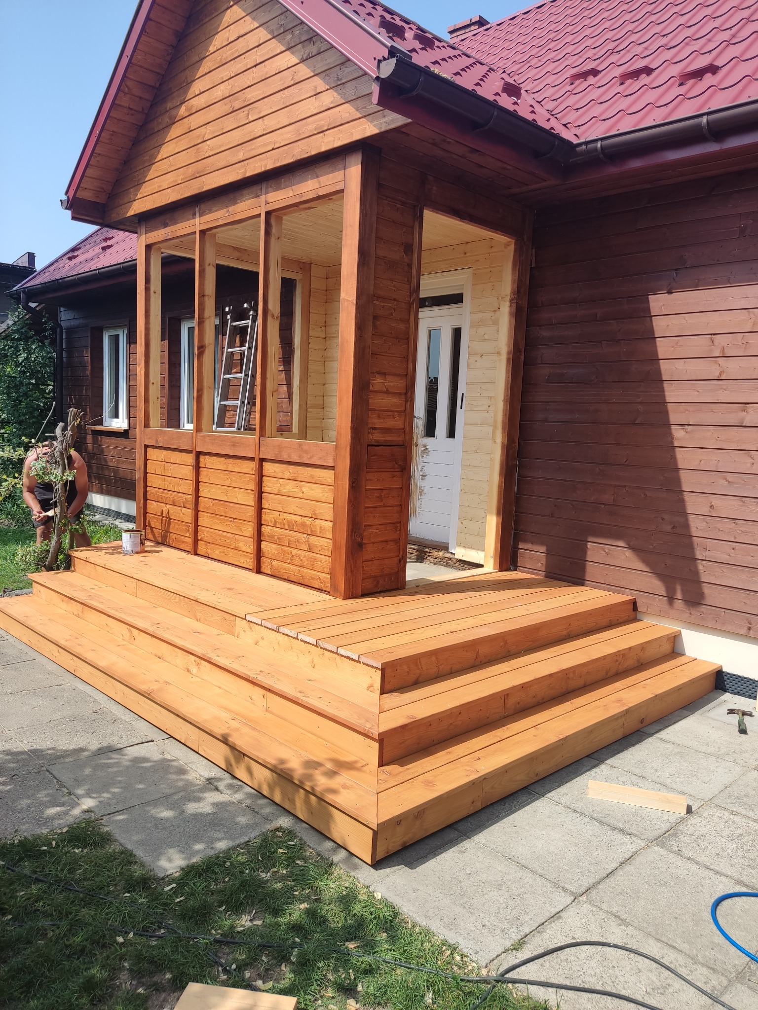 Nowo wybudowany, drewniany taras z wejściem do domu, świeżo pomalowany na brązowo, z widoczną drabiną wewnątrz i osobą malującą detale na zewnątrz. Widoczna puszka z farbą i narzędzia.