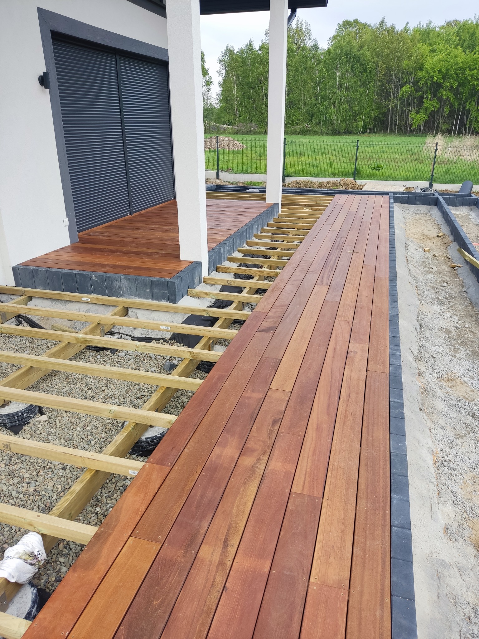 Montaż tarasu drewnianego z egzotycznego drewna: widoczna konstrukcja nośna z belek na podsypce żwirowej, częściowo ułożone deski tarasowe, obrzeże z kostki brukowej, tło z zielenią