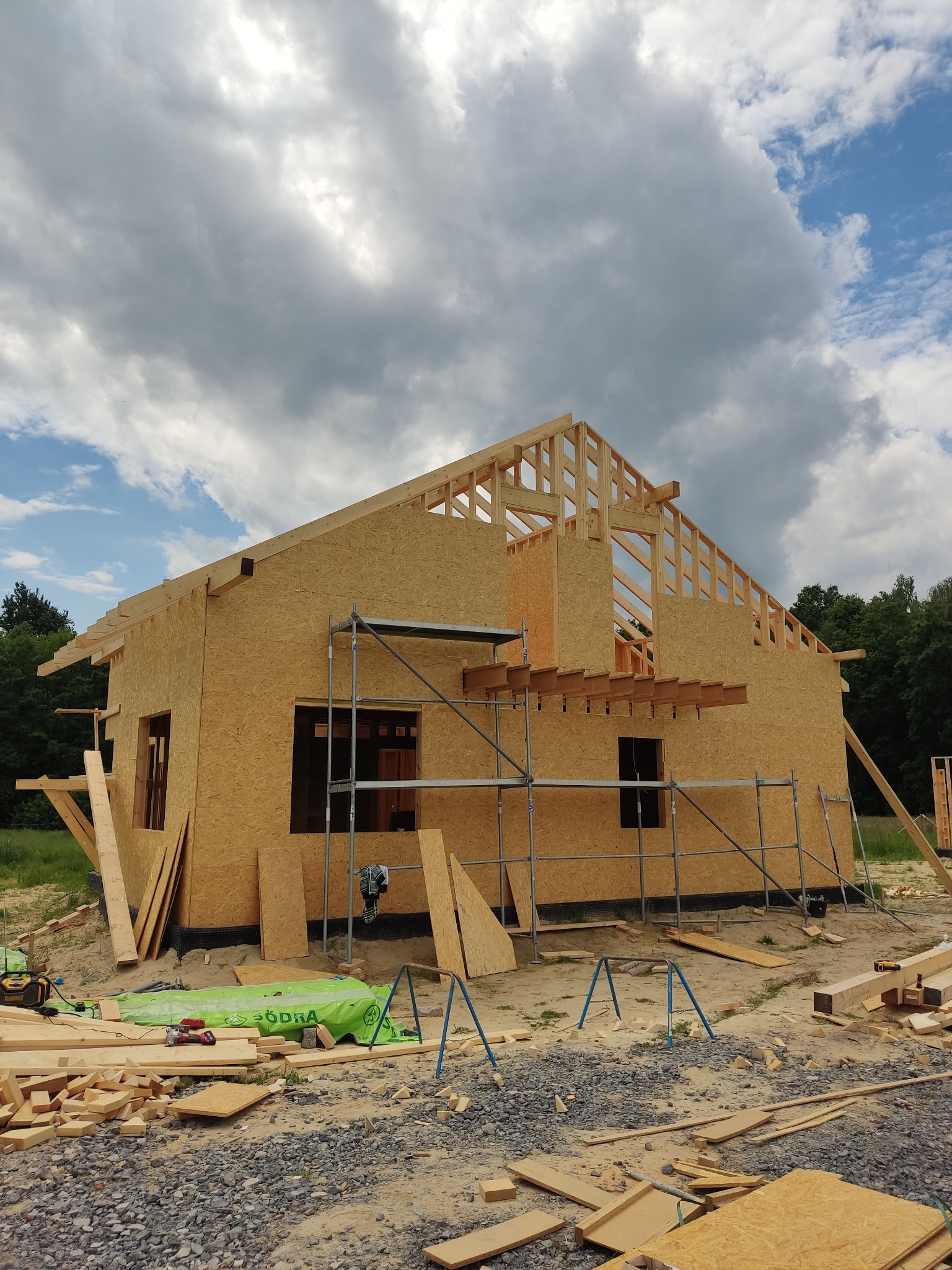 Konstrukcja domu w trakcie budowy, widoczne ściany z płyt OSB, drewniany szkielet dachu, rusztowanie, oraz materiały budowlane na ziemi, pochmurne niebo w tle.