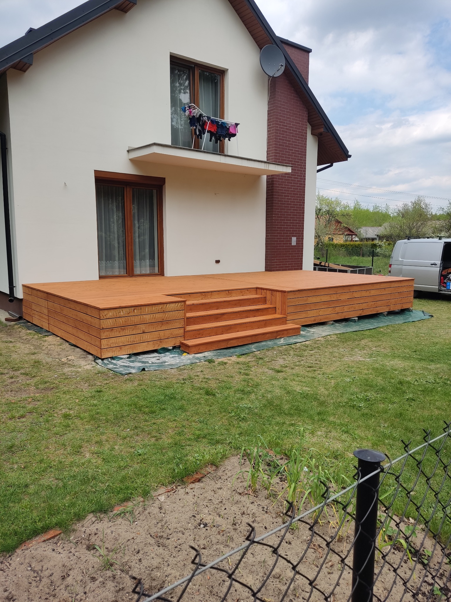 Nowy, drewniany taras z niskimi schodami, przylegający do białego domu z ceglanym kominem i suszarką balkonową.