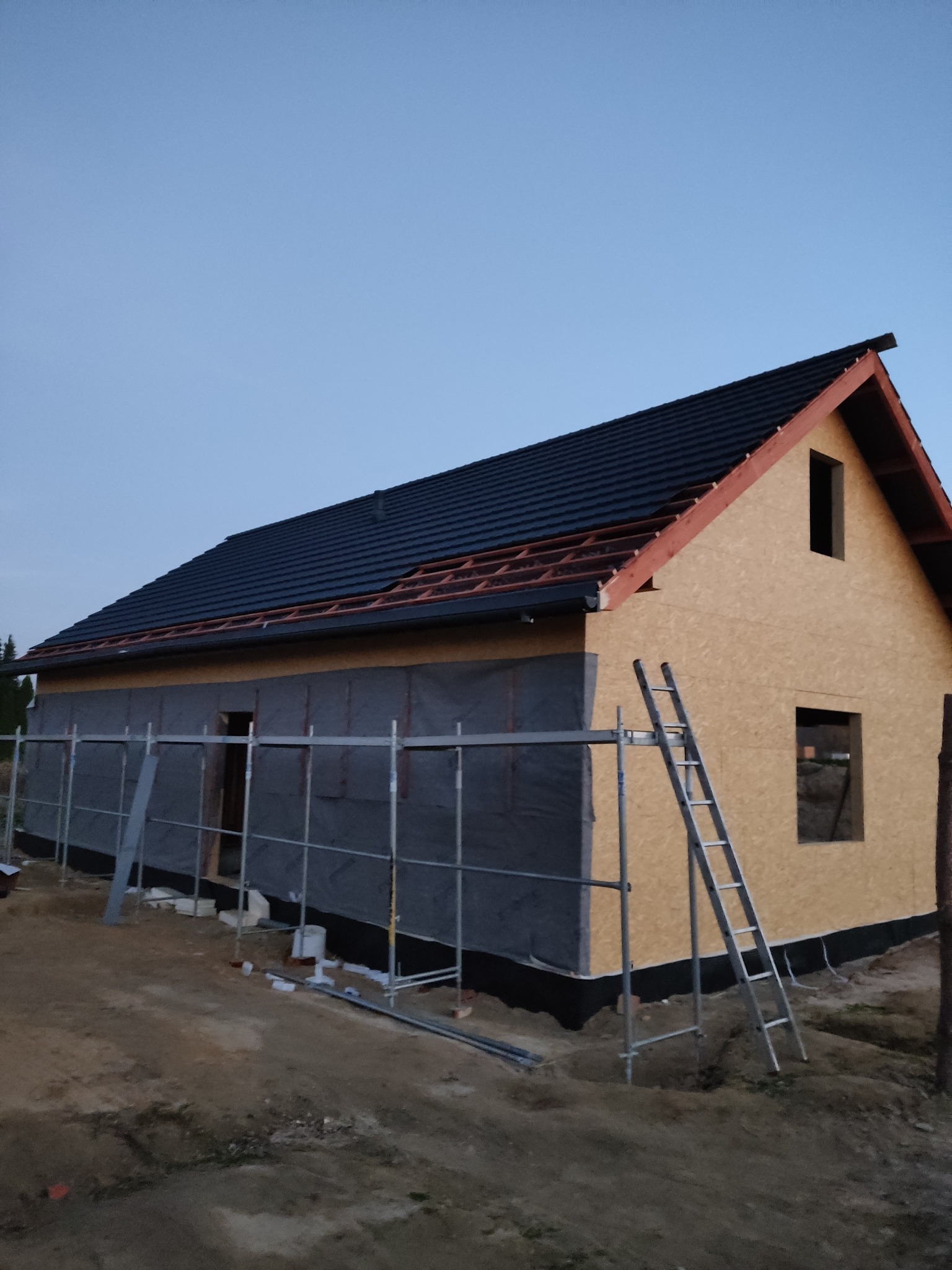 Budowa domu w trakcie: surowa konstrukcja z płyt OSB, czarne pokrycie dachu częściowo ułożone, rusztowania i drabina przy ścianie, widoczne otwory okienne, przygotowanie do ocieplenia.