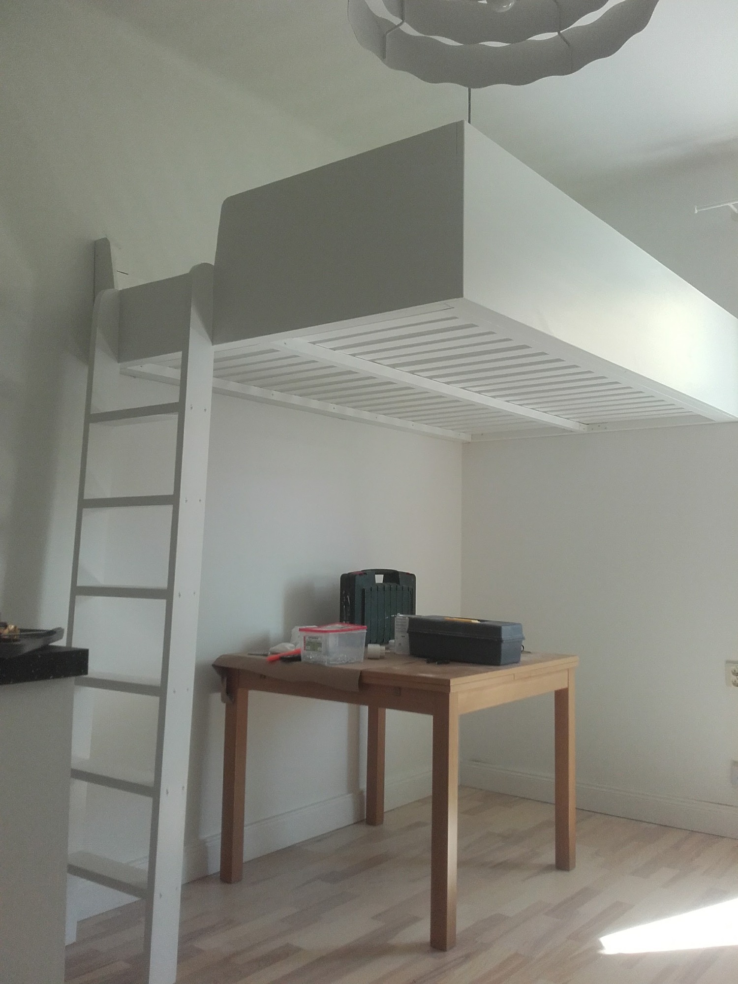 Pokój z białym łóżkiem na antresoli z drabinką, pod spodem drewniany stół z narzędziami w pudełkach, oświetlony lampą sufitową.