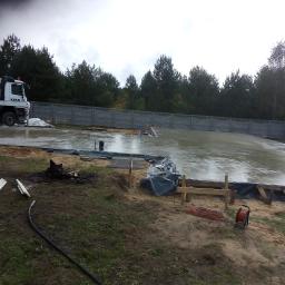 Damian Pichliński - Wylany, mokry beton na przygotowanym fundamencie pod budowę, widoczny żuraw CIFA, szary płot w tle i teren budowy z elementami szalunku.