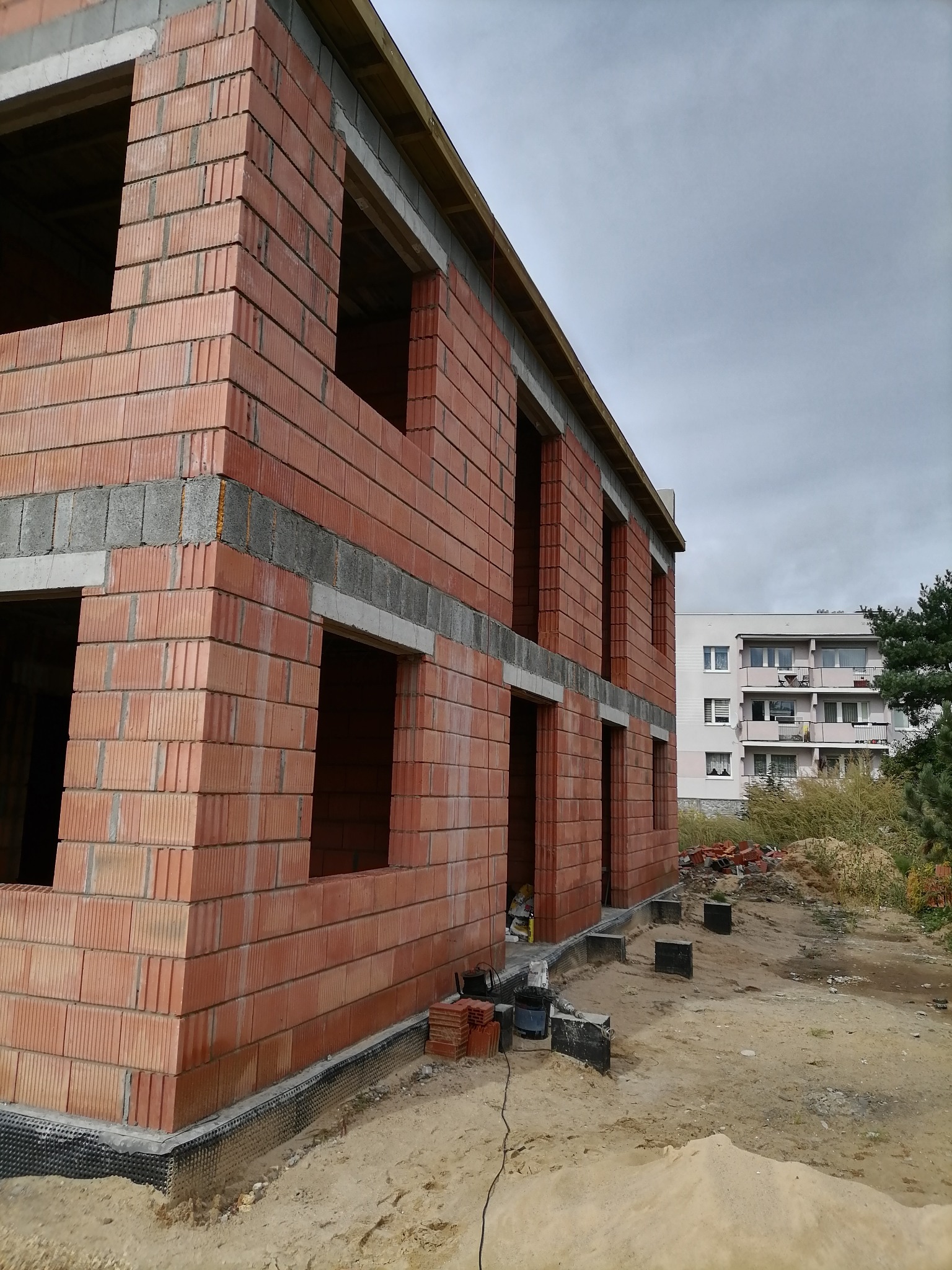 Dwukondygnacyjny dom murowany w trakcie budowy, widoczne ściany z czerwonej cegły, otwory okienne i drzwiowe, szary betonowy wieniec, fundament zabezpieczony folią kubełkową, w tle blok mieszkalny...