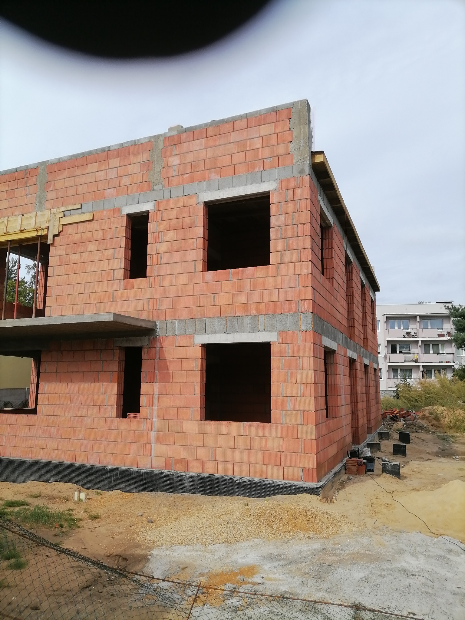 Dwukondygnacyjny budynek w trakcie budowy, wzniesiony z czerwonej cegły kratówki, z widocznymi otworami okiennymi i betonowymi nadprożami, osadzony na fundamencie zabezpieczonym czarną folią.