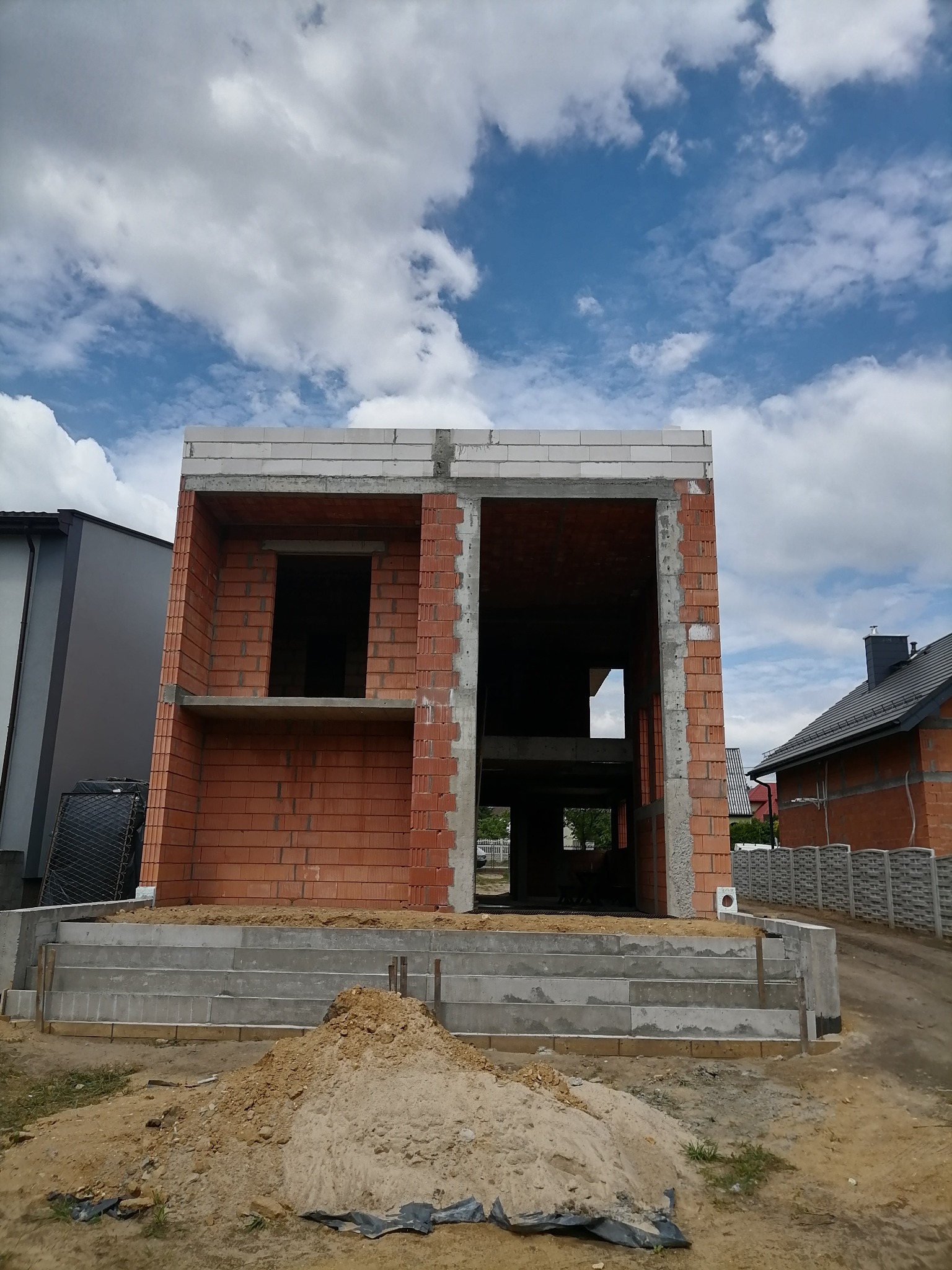 Surowy stan dwukondygnacyjnego domu z czerwonej cegły i betonu komórkowego, widok z zewnątrz na wczesnym etapie budowy, z widocznym fundamentem i stertą ziemi na pierwszym planie.