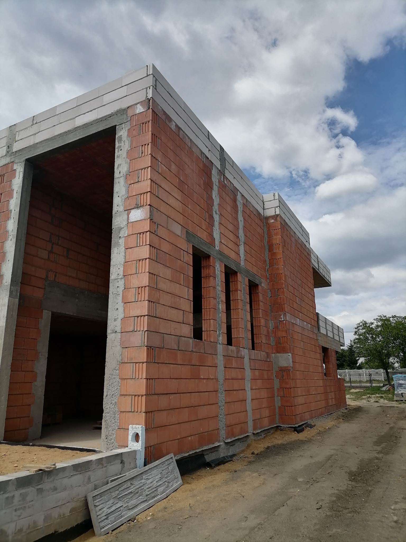 Surowy stan budynku z czerwonej cegły szczelinowej, widoczne otwory okienne i betonowe nadproża, ściany w trakcie wznoszenia, fragment fundamentu i nieba z chmurami w tle.