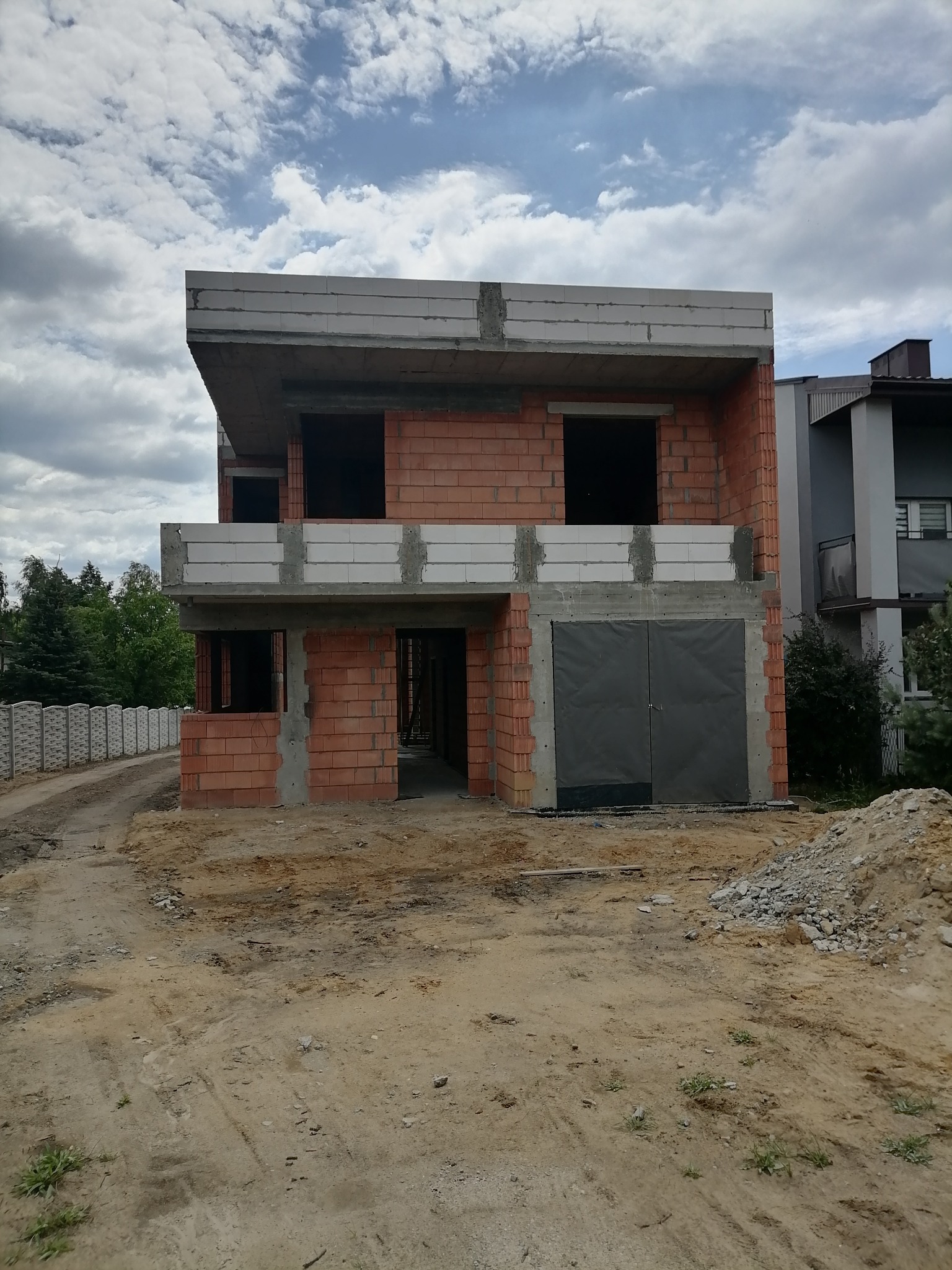 Dwukondygnacyjny budynek w stanie surowym otwartym, wzniesiony z czerwonej cegły i białych bloczków, z prowizoryczną bramą garażową, na tle pochmurnego nieba i sąsiedniego domu.