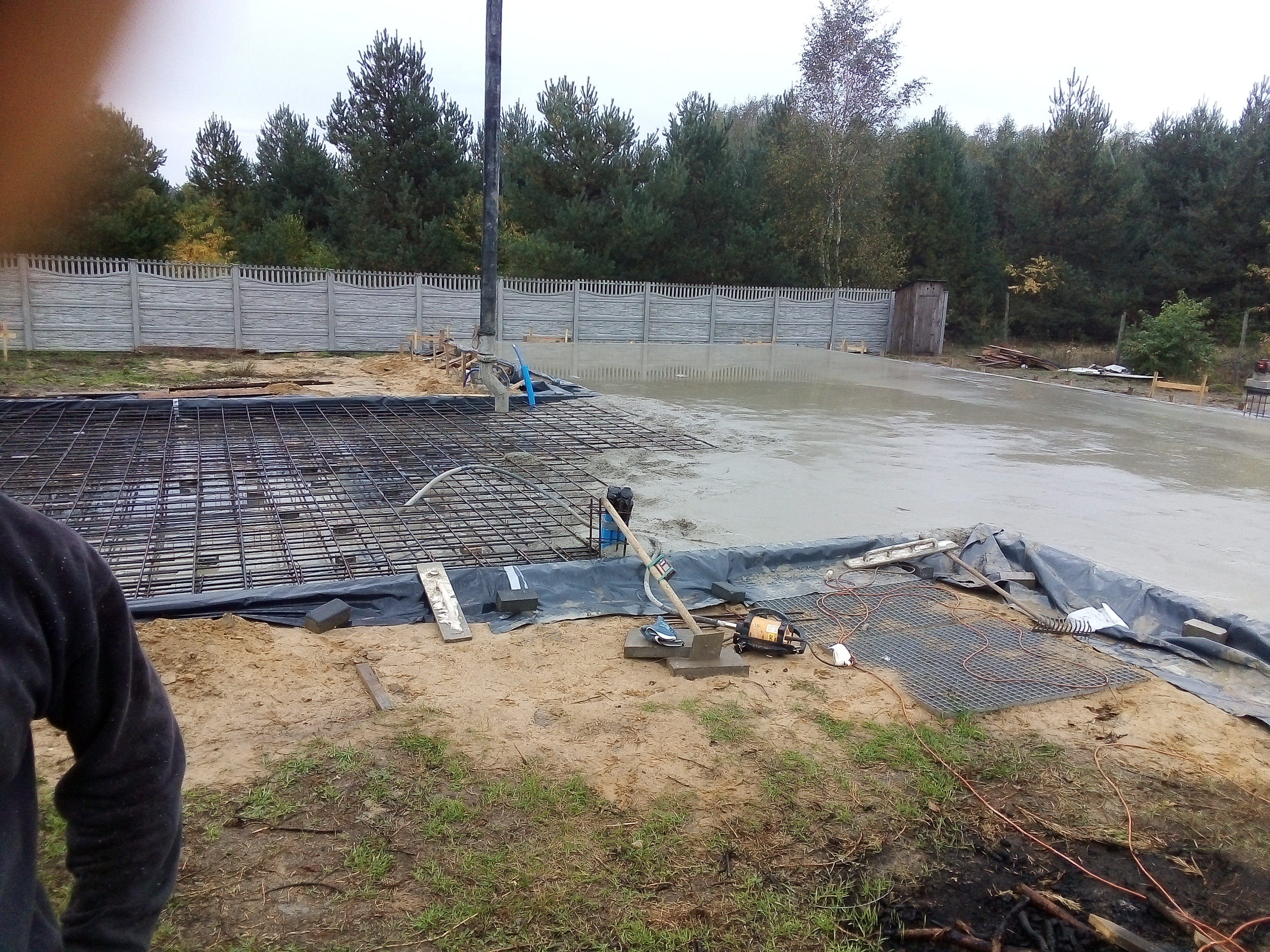 Wylewanie betonu na przygotowany fundament z siatką zbrojeniową, widoczna pompa do betonu, teren zabezpieczony folią, w tle ogrodzenie panelowe i drzewa.
