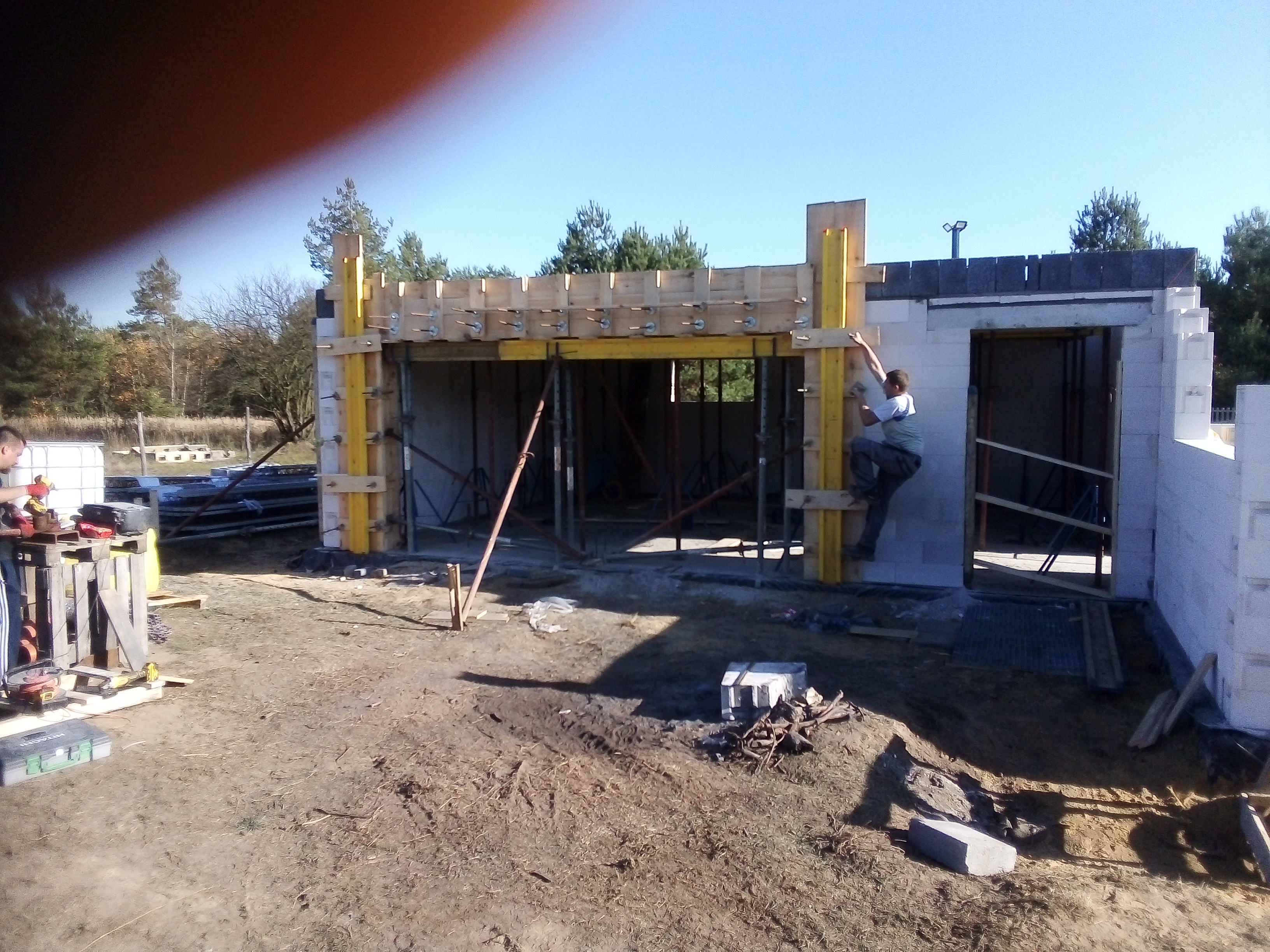 Wznoszenie ścian parterowego budynku z bloczków, z widocznym szalunkiem systemowym w kolorze żółtym, pracownik wspina się po szalunku, w tle las i błękitne niebo.