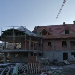 Budowa domu z czerwonej cegły z widoczną konstrukcją dachu i częściowo ułożoną czerwoną dachówką, pracownicy na rusztowaniach, w tle góry i żuraw budowlany.