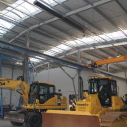 Wnętrze hali z konstrukcją stalową dachu, oświetleniem i suwnicą, gdzie stoją dwie żółte maszyny budowlane marki Komatsu: koparka kołowa PW160 i spycharka 61PX-15.