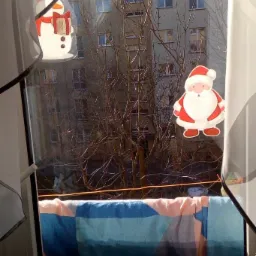 Widok z okna na blok mieszkalny z nagimi drzewami, ozdobionego naklejkami Świętego Mikołaja i bałwana, z rozwieszonym praniem na sznurku.