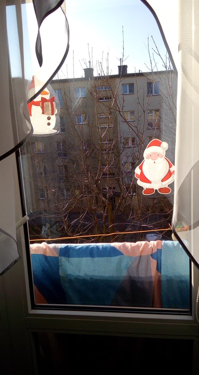 Widok z okna na blok mieszkalny z nagimi drzewami, ozdobionego naklejkami Świętego Mikołaja i bałwana, z rozwieszonym praniem na sznurku.