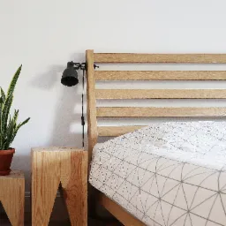 Minimalistyczna sypialnia z drewnianym łóżkiem, geometryczną pościelą, lampką nocną i rośliną doniczkową na drewnianych stolikach nocnych.