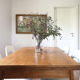 AGNIESZKA LISEK architekt wnętrz - Stół z litego drewna z bukietem polnych kwiatów w szklanym wazonie, białe krzesła, obraz w złotej ramie i grzejnik w tle, minimalistyczna aranżacja jadalni w Kaliszu.