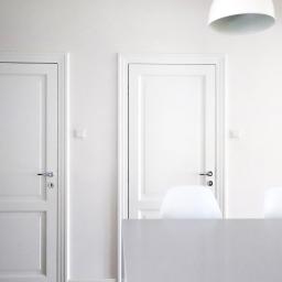 AGNIESZKA LISEK architekt wnętrz - Minimalistyczne wnętrze: dwie białe, klasyczne drzwi z srebrnymi klamkami na białej ścianie, białe krzesła i lampa sufitowa w tle, utrzymane w jednolitej, jasnej kolorystyce.