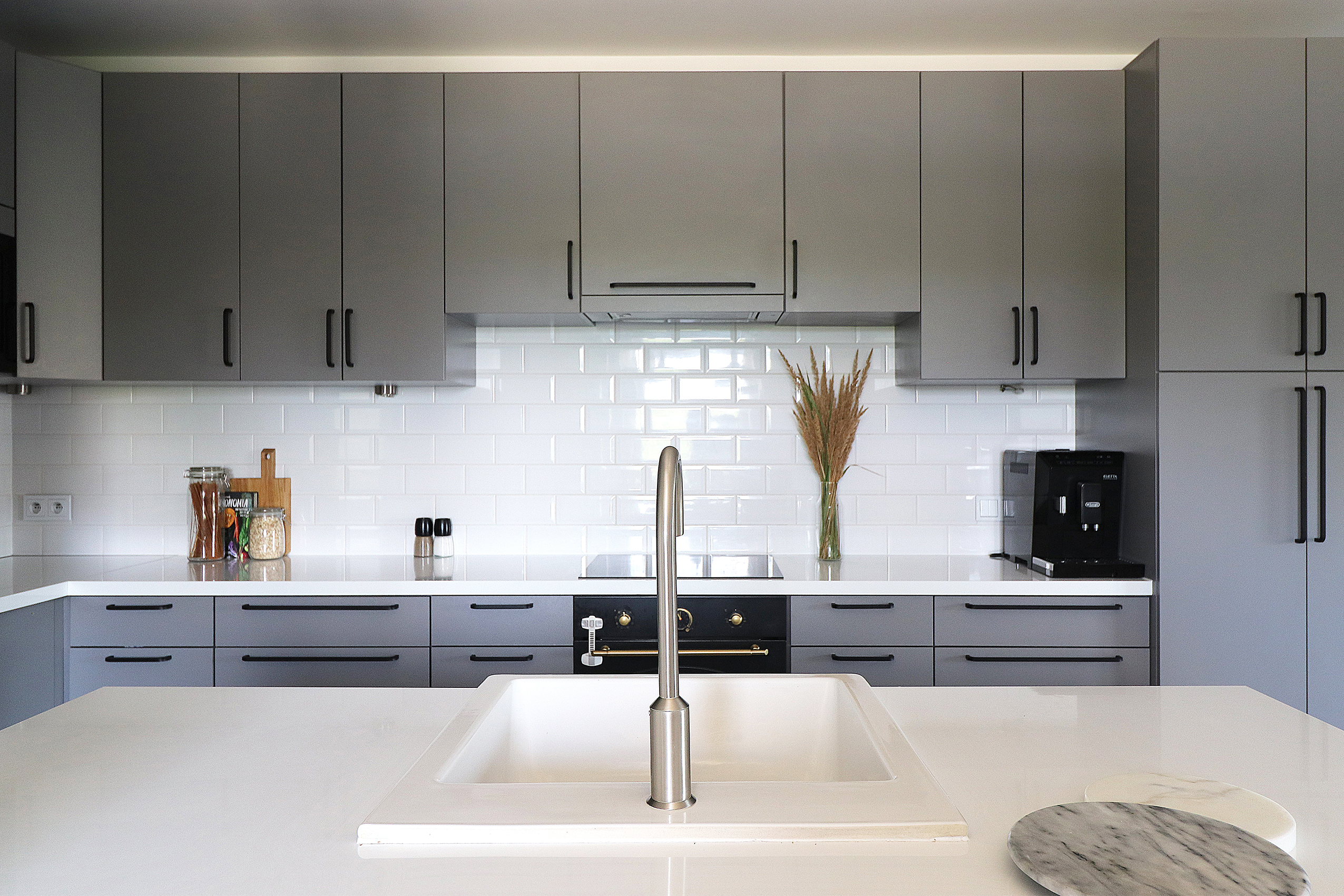 Nowoczesna kuchnia w szarościach z białym blatem i płytkami typu metro; widoczny zlewozmywak z baterią, płyta indukcyjna, ekspres do kawy i dekoracyjne trawy w wazonie.