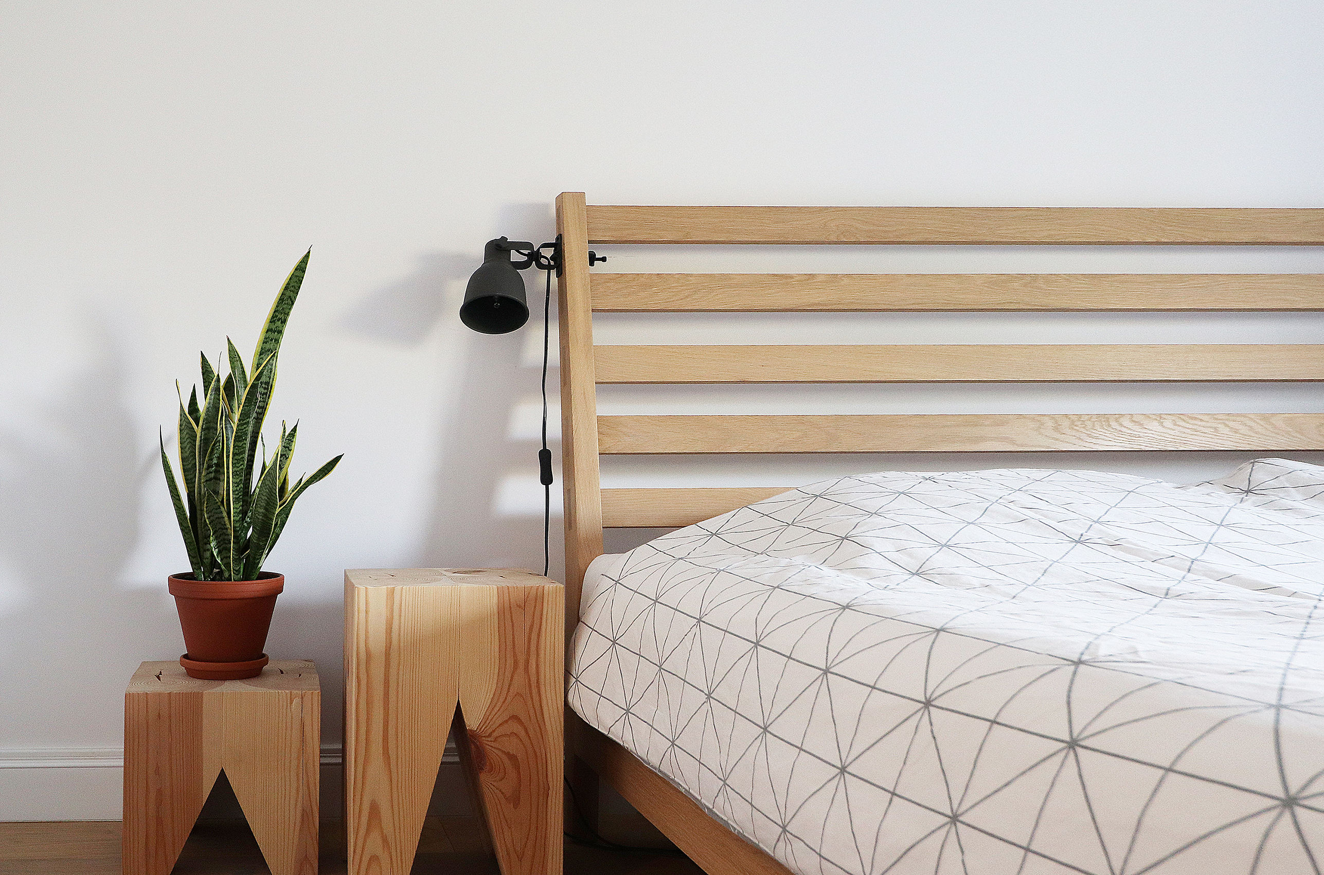 Minimalistyczna sypialnia z drewnianym łóżkiem, geometryczną pościelą, lampką nocną i rośliną doniczkową na drewnianych stolikach nocnych.