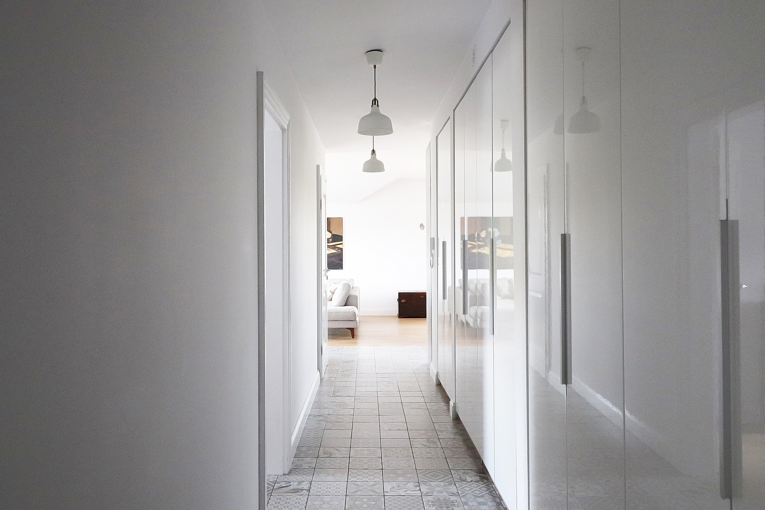 Długi, jasny korytarz z błyszczącą białą szafą w zabudowie, wzorzystą podłogą z płytek i widokiem na salon z szarą sofą i ciemną skrzynią.