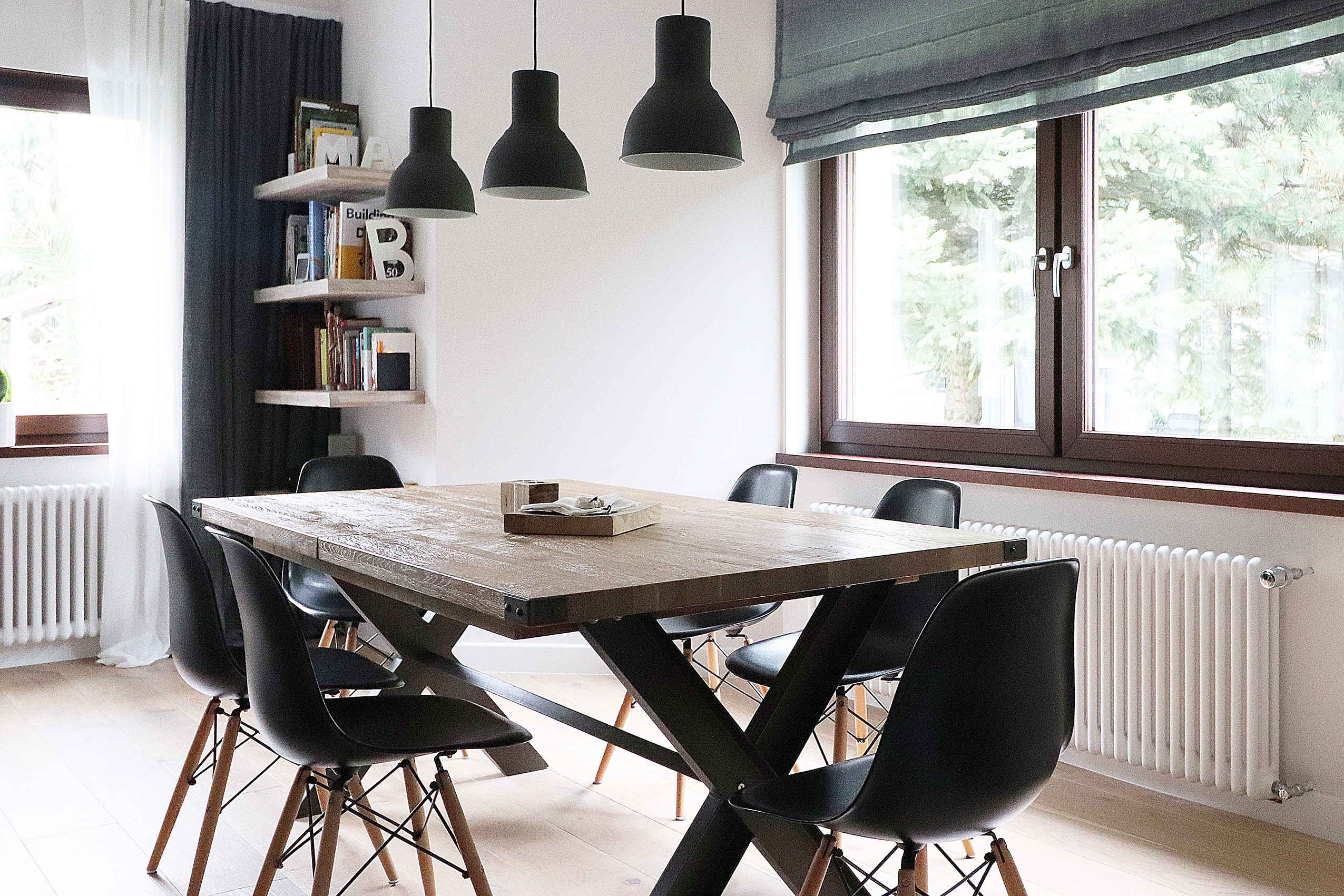 Jadalnia w nowoczesnym stylu z drewnianym stołem na metalowej podstawie w kształcie litery X, czarnymi krzesłami Eames, trzema wiszącymi lampami w kształcie dzwonów, półkami z książkami i roletą...