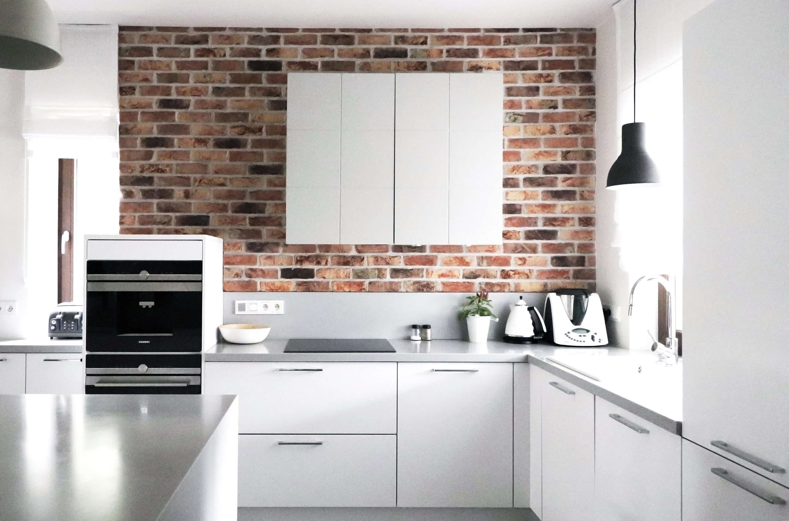Jasna kuchnia z białymi szafkami i blatem, ceglana ściana w tle, nowoczesne oświetlenie wiszące, sprzęty AGD na blacie, roślina w doniczce.