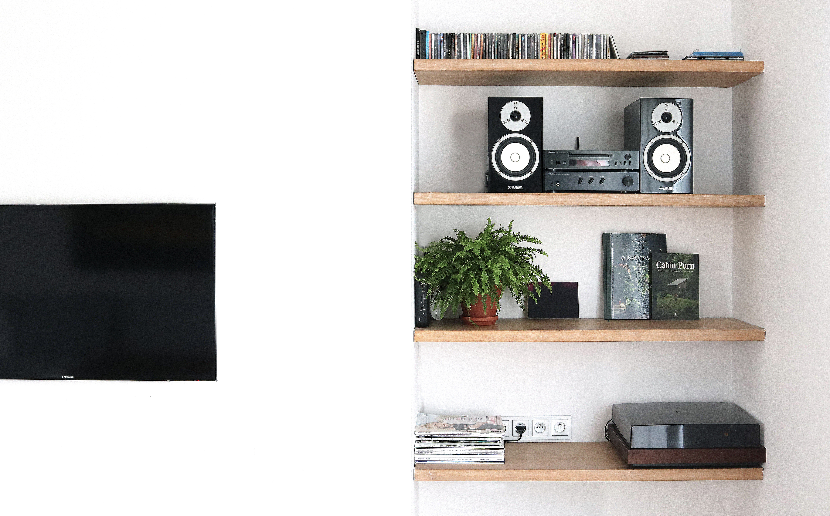 Telewizor na ścianie, półki z jasnego drewna z głośnikami Yamaha, wzmacniaczem, płytami CD, paprotką w doniczce, książkami i gramofonem, minimalistyczny salon.