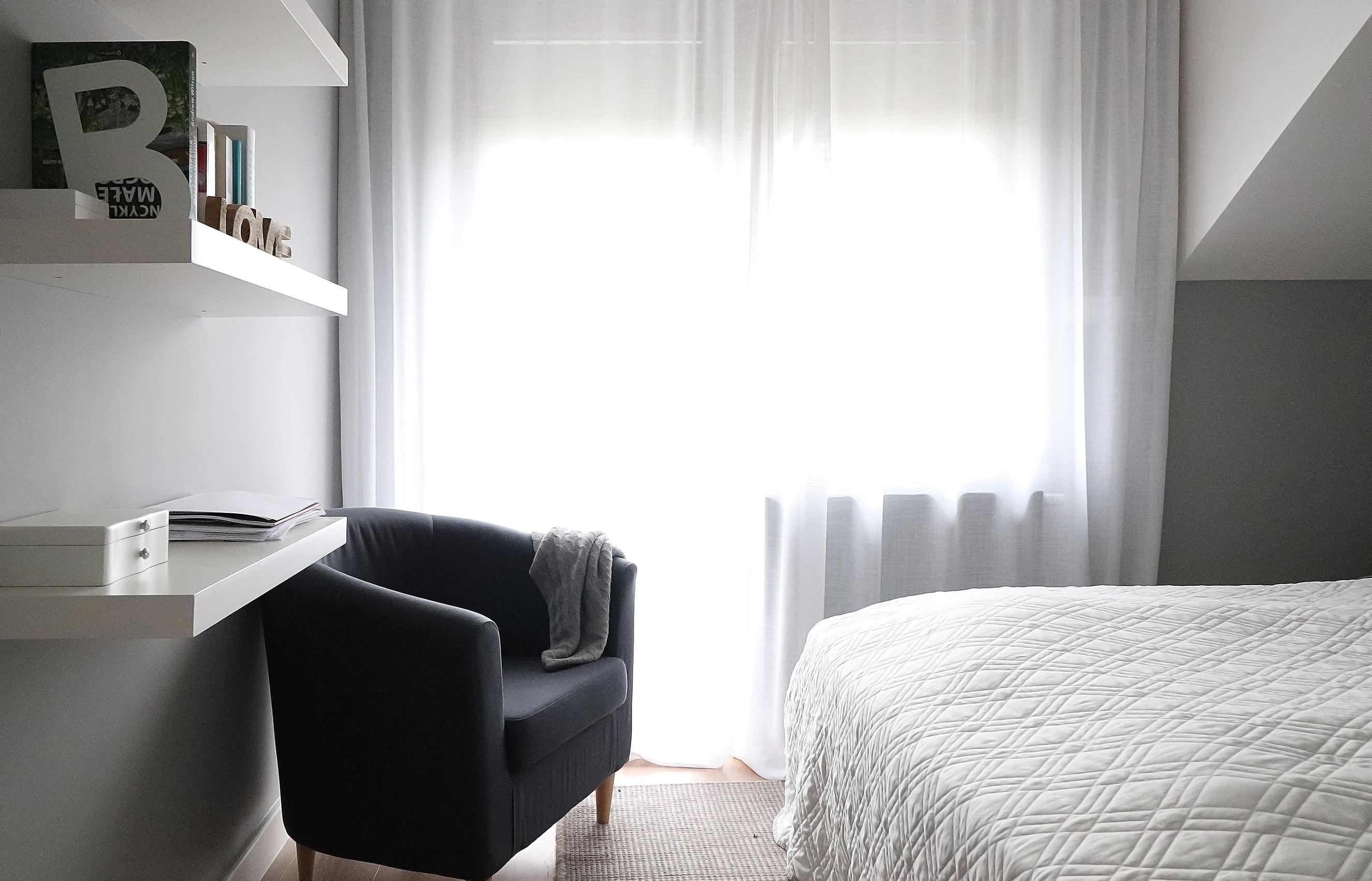 Jasne wnętrze sypialni na poddaszu z białymi zasłonami, szarym fotelem z narzutą, biurkiem z półkami i białym łóżkiem z pikowaną narzutą.