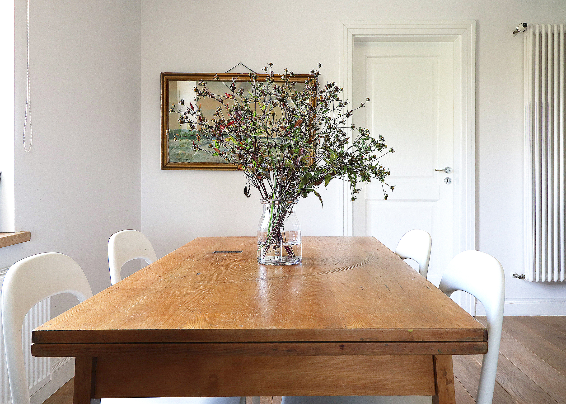 Stół z litego drewna z bukietem polnych kwiatów w szklanym wazonie, białe krzesła, obraz w złotej ramie i grzejnik w tle, minimalistyczna aranżacja jadalni w Kaliszu.