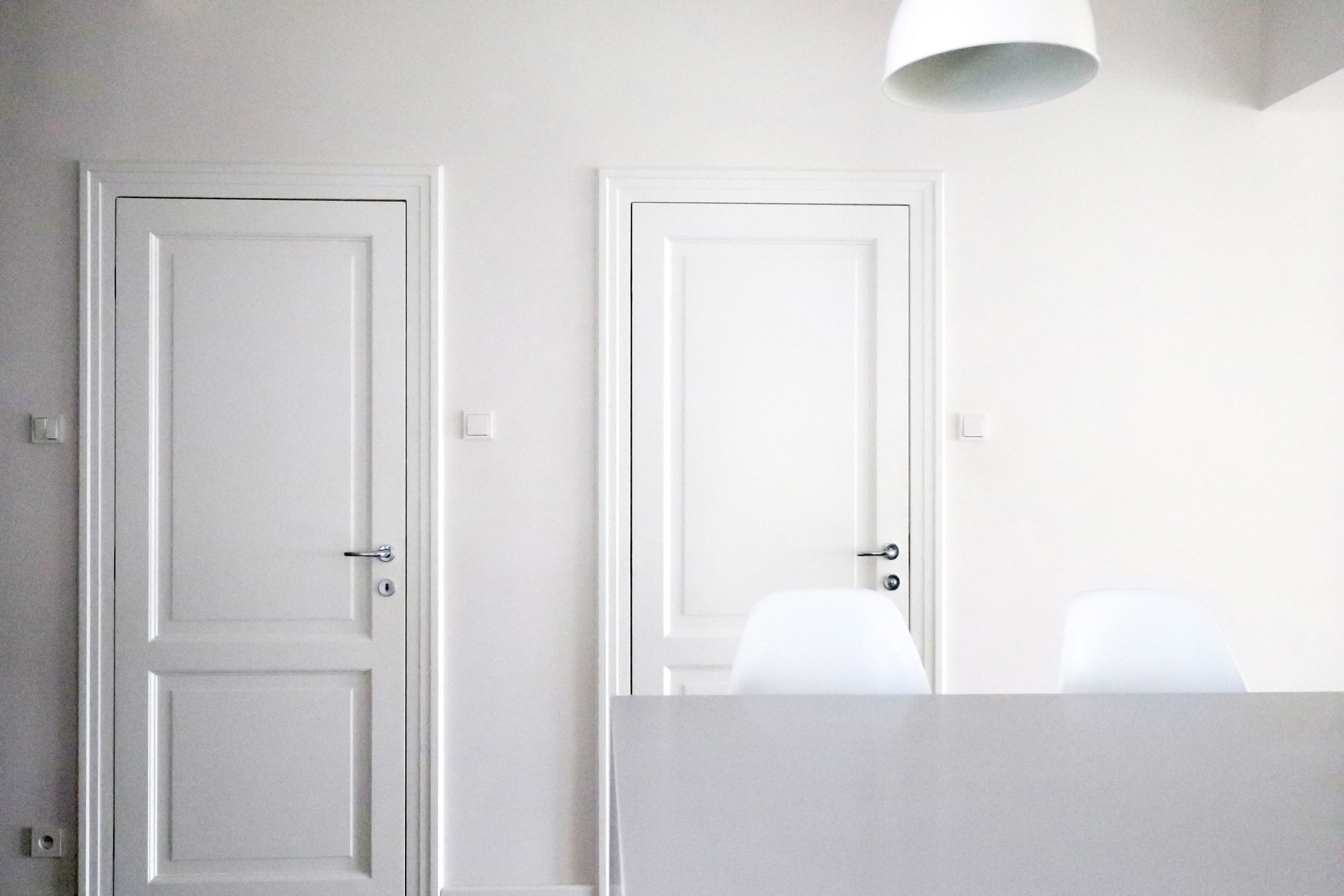 Minimalistyczne wnętrze: dwie białe, klasyczne drzwi z srebrnymi klamkami na białej ścianie, białe krzesła i lampa sufitowa w tle, utrzymane w jednolitej, jasnej kolorystyce.