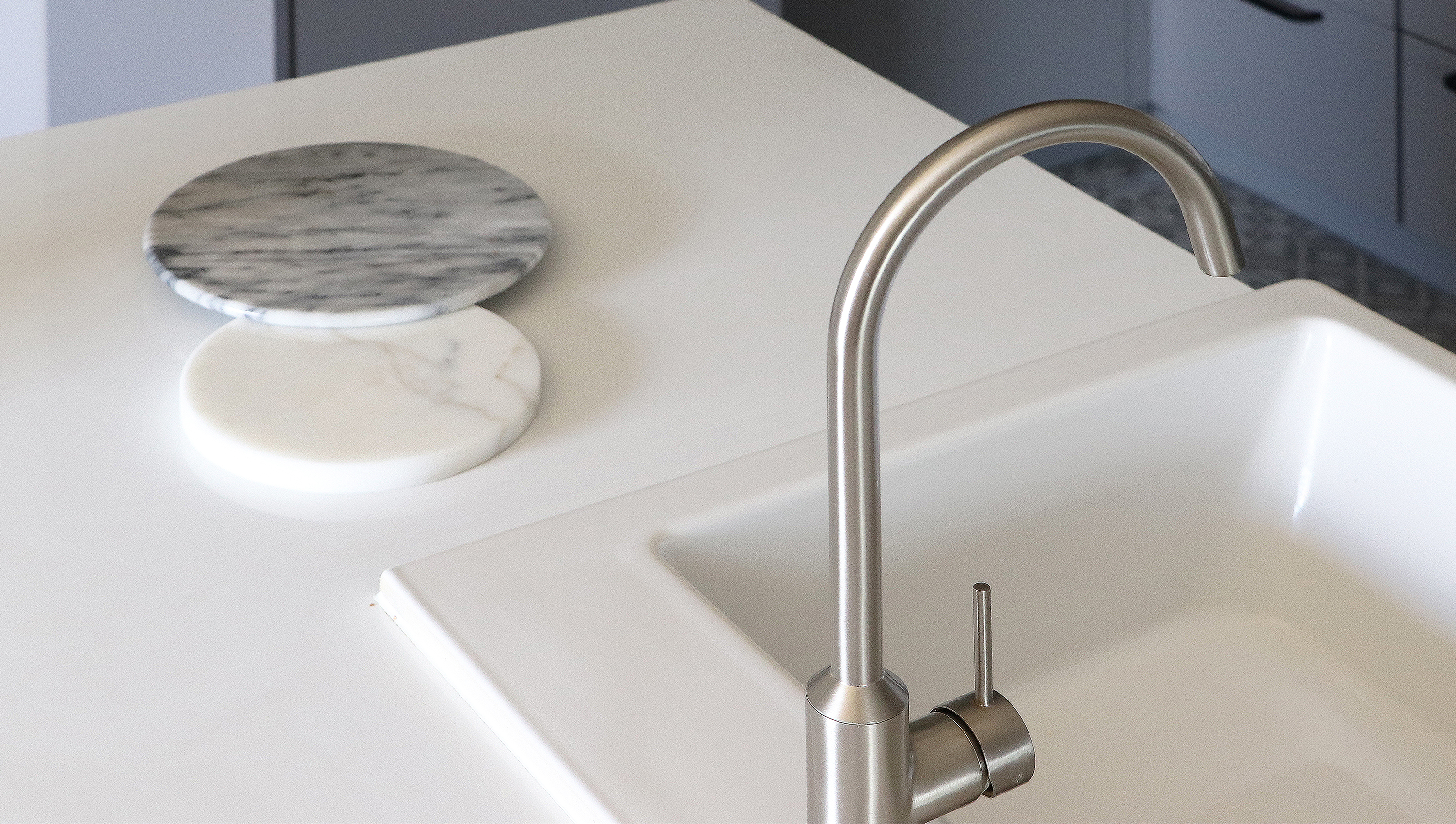 Biała, prostokątna umywalka z nowoczesną, metalową baterią o łukowatym kształcie, obok leżą dwa okrągłe podkładki z marmuru, jedna biała i jedna szara.