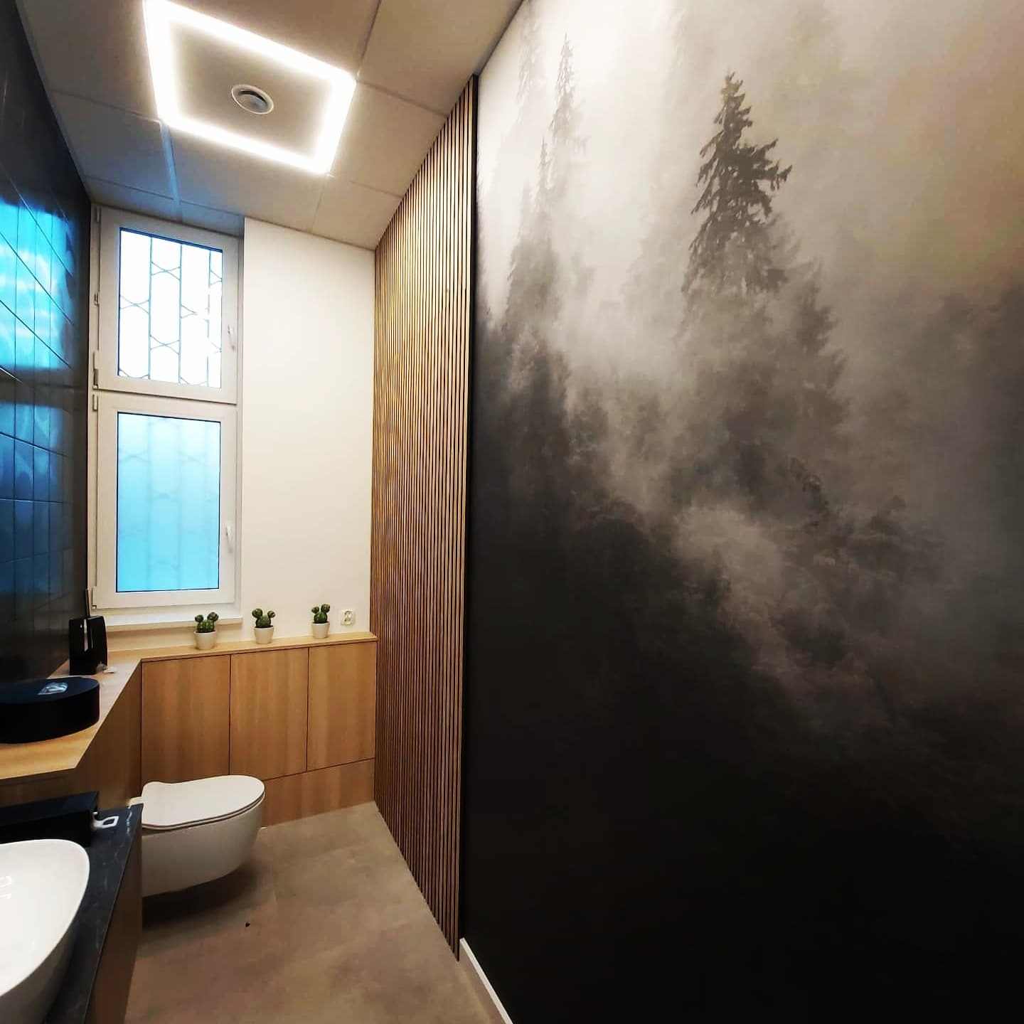 Nowoczesna łazienka z zabudową meblową z jasnego drewna, toaletą podwieszaną, ścianą z drewnianych lameli i fototapetą z motywem mglistego lasu.