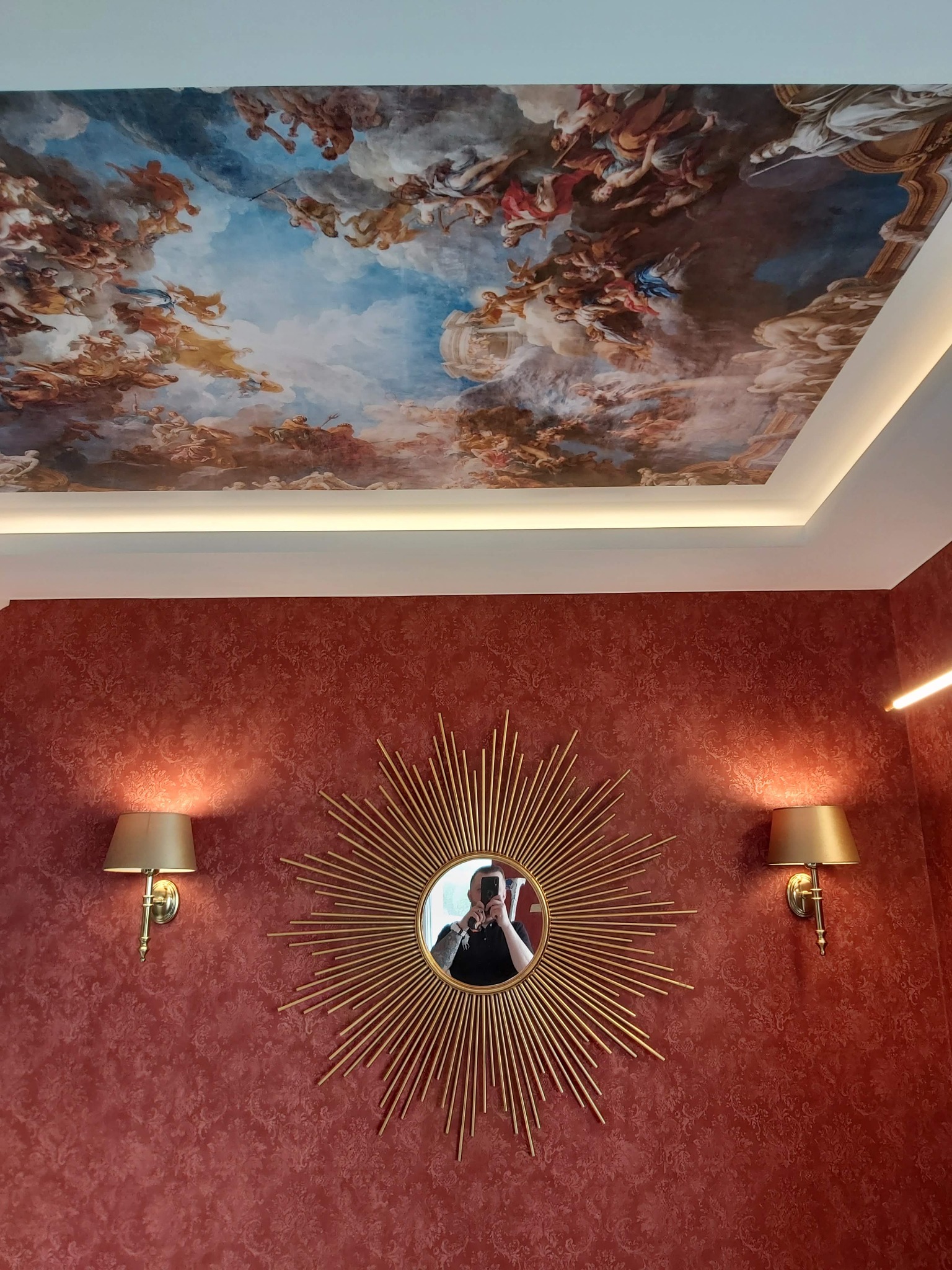 Czerwona ściana z wzorem, lustro w złotej ramie w kształcie słońca i dwa kinkiety, sufit z malowidłem przedstawiającym sceny mitologiczne, oświetlony listwą LED.