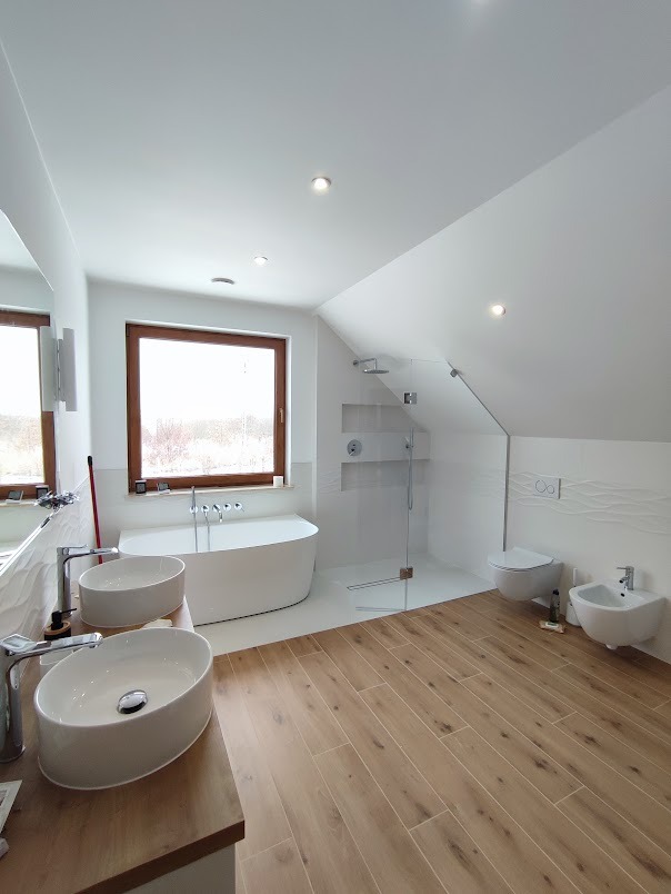 Jasna łazienka na poddaszu z drewnopodobną podłogą, owalną wanną wolnostojącą pod oknem, dwoma umywalkami nablatowymi i kabiną prysznicową typu walk-in.