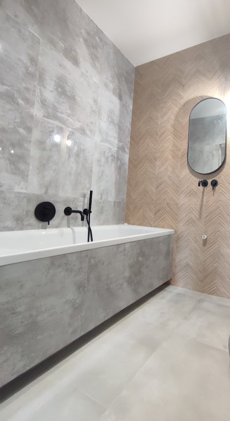 Nowoczesna łazienka z wanną obudowaną szarymi płytami imitującymi beton, czarna armatura, lustro w owalnej ramie i ściana wyłożona płytkami w jodełkę.