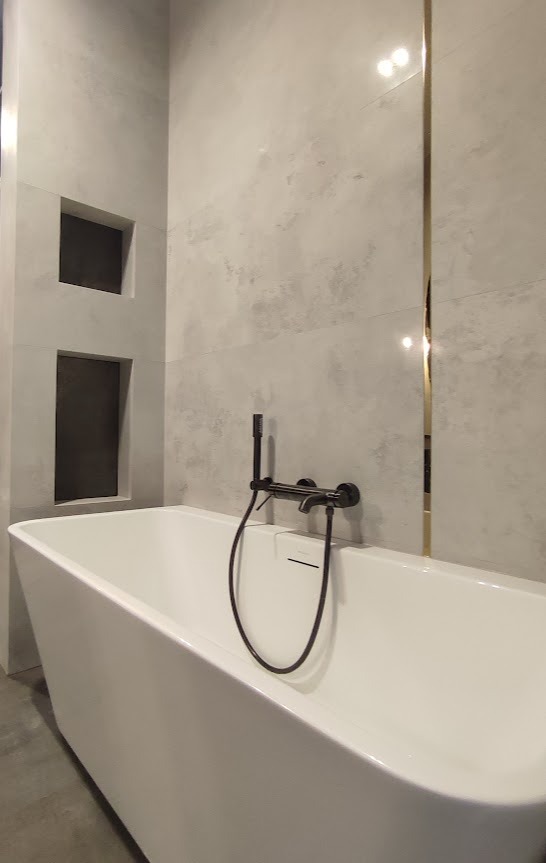 Nowoczesna łazienka z białą, wolnostojącą wanną, czarną armaturą i szarymi płytkami imitującymi beton, z wnękami w ścianie.