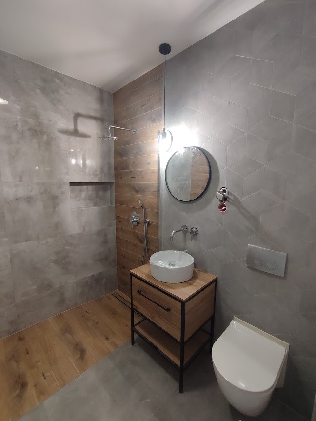 Nowoczesna łazienka z drewnianymi akcentami: kabina prysznicowa typu walk-in z szarymi płytkami imitującymi beton, umywalka nablatowa na drewnianej szafce z czarnymi metalowymi nogami, okrągłe...