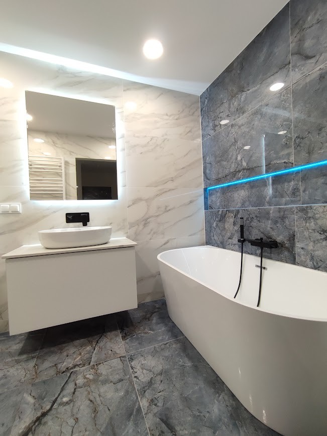 Nowoczesna łazienka z marmurowymi płytkami, podświetlanym lustrem nad wiszącą szafką z umywalką i wolnostojącą wanną z czarną armaturą.