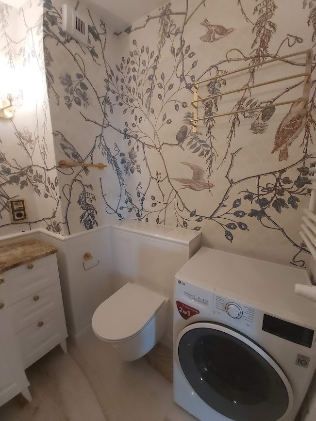 Nowoczesna łazienka z toaletą podwieszaną, pralką LG i szafką umywalkową w stylu retro, wyróżniająca się tapetą w motywy roślinne i ptasie w kolorach szarości i beżu.