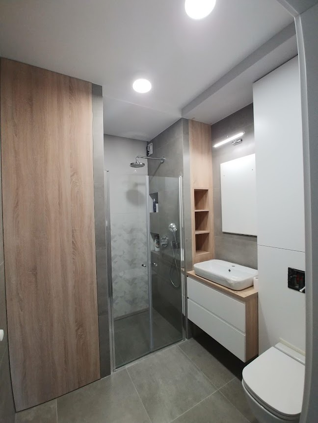 Nowoczesna łazienka z kabiną prysznicową typu walk-in, umywalką nablatową na szafce z drewnianym blatem oraz wiszącą toaletą, wykończona szarymi płytkami i drewnopodobnymi panelami ściennymi.