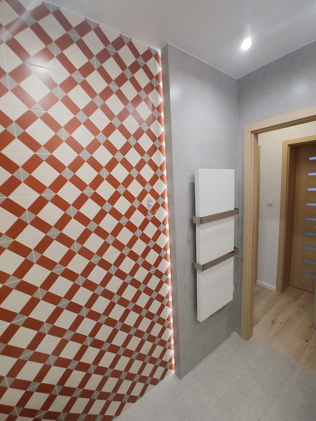 Nowoczesna łazienka z geometryczną ścianą w romby w odcieniach bieli i terakoty, biały grzejnik drabinkowy na szarej ścianie, widok na drewniane drzwi w tle.