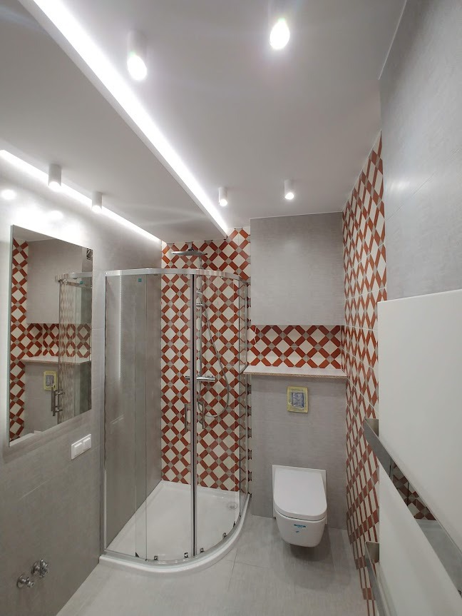 Nowoczesna łazienka z narożną kabiną prysznicową ze srebrnymi profilami, toaletą wiszącą, szarą glazurą i dekoracyjną ścianą w geometryczne wzory w odcieniach czerwieni i bieli.