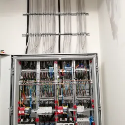 Otwarta szafa elektryczna z licznymi przewodami ułożonymi w korytkach, widoczne zabezpieczenia, styczniki i moduły, białe ściany w tle.