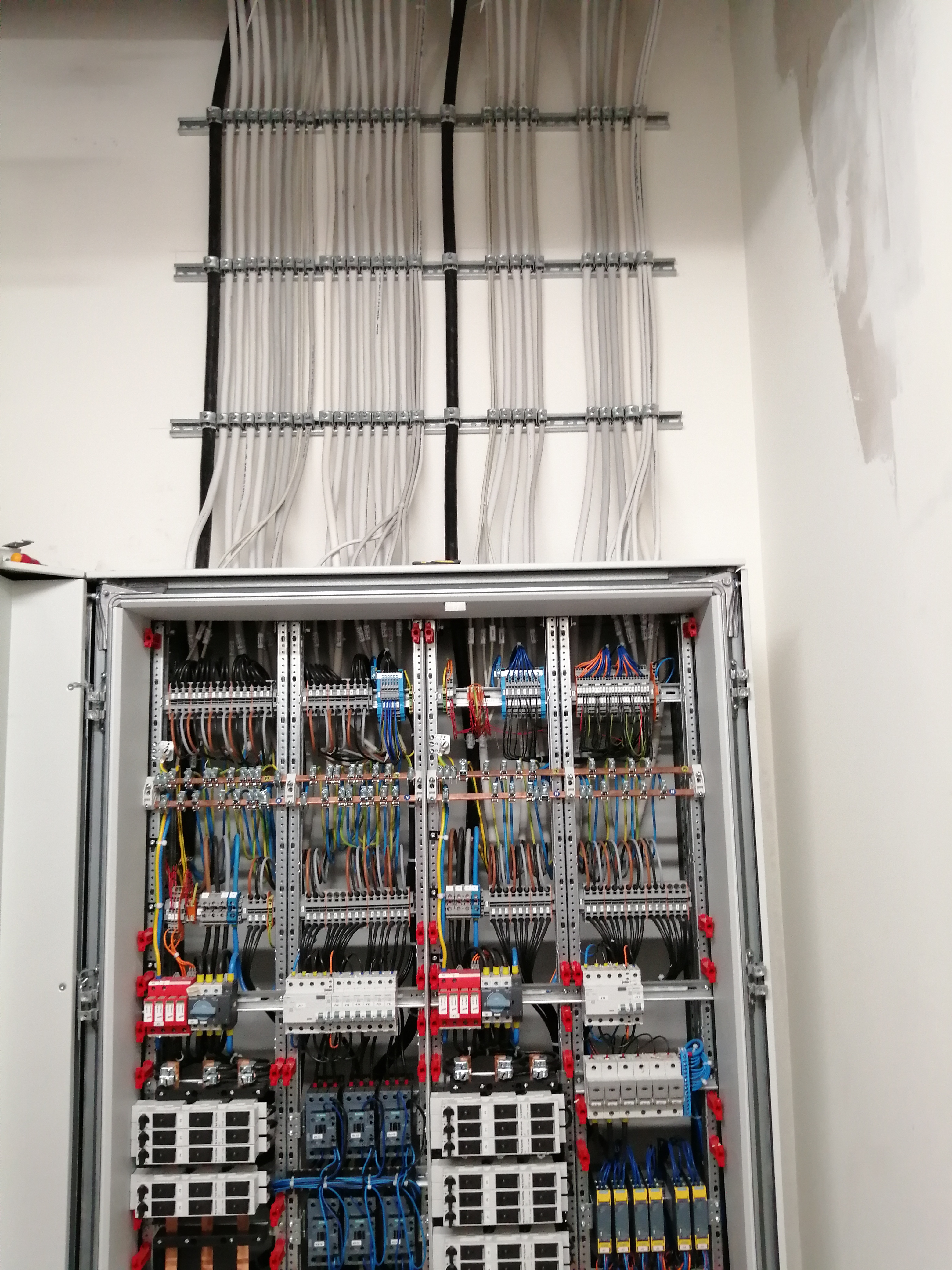 Otwarta szafa elektryczna z licznymi przewodami ułożonymi w korytkach, widoczne zabezpieczenia, styczniki i moduły, białe ściany w tle.