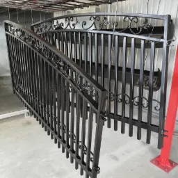 Metalowa balustrada schodowa z ozdobnymi elementami, świeżo po nałożeniu czarnej powłoki, zawieszona na specjalnej konstrukcji w hali produkcyjnej.