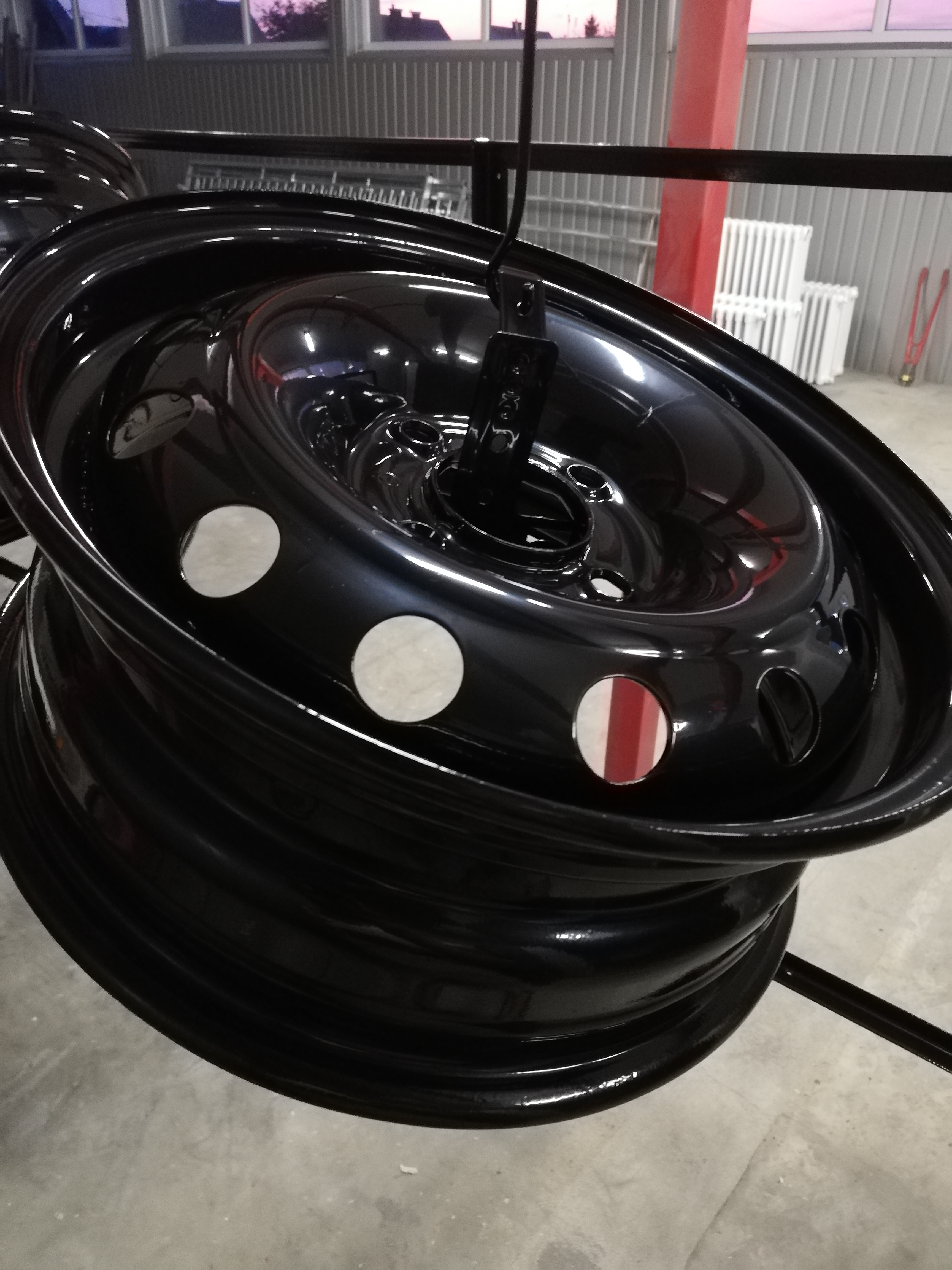 Błyszcząca, czarna felga samochodowa zawieszona na haku w warsztacie, widoczne białe i czerwone okrągłe naklejki maskujące otwory montażowe.