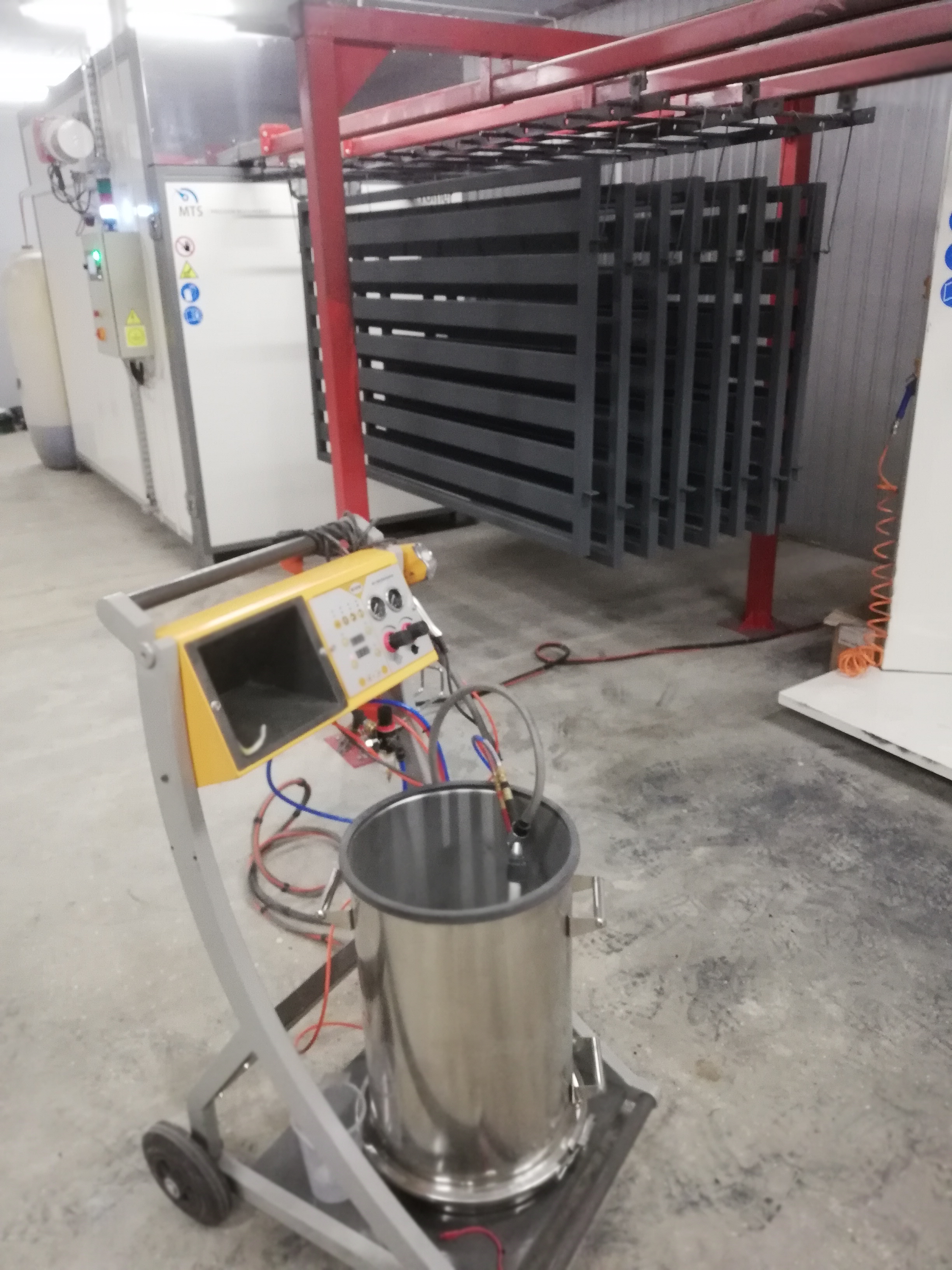 Elementy metalowe po malowaniu proszkowym, zawieszone na wózku transportowym, widoczna maszyna do malowania proszkowego z żółtym panelem sterowania i stalowym zbiornikiem na proszek.