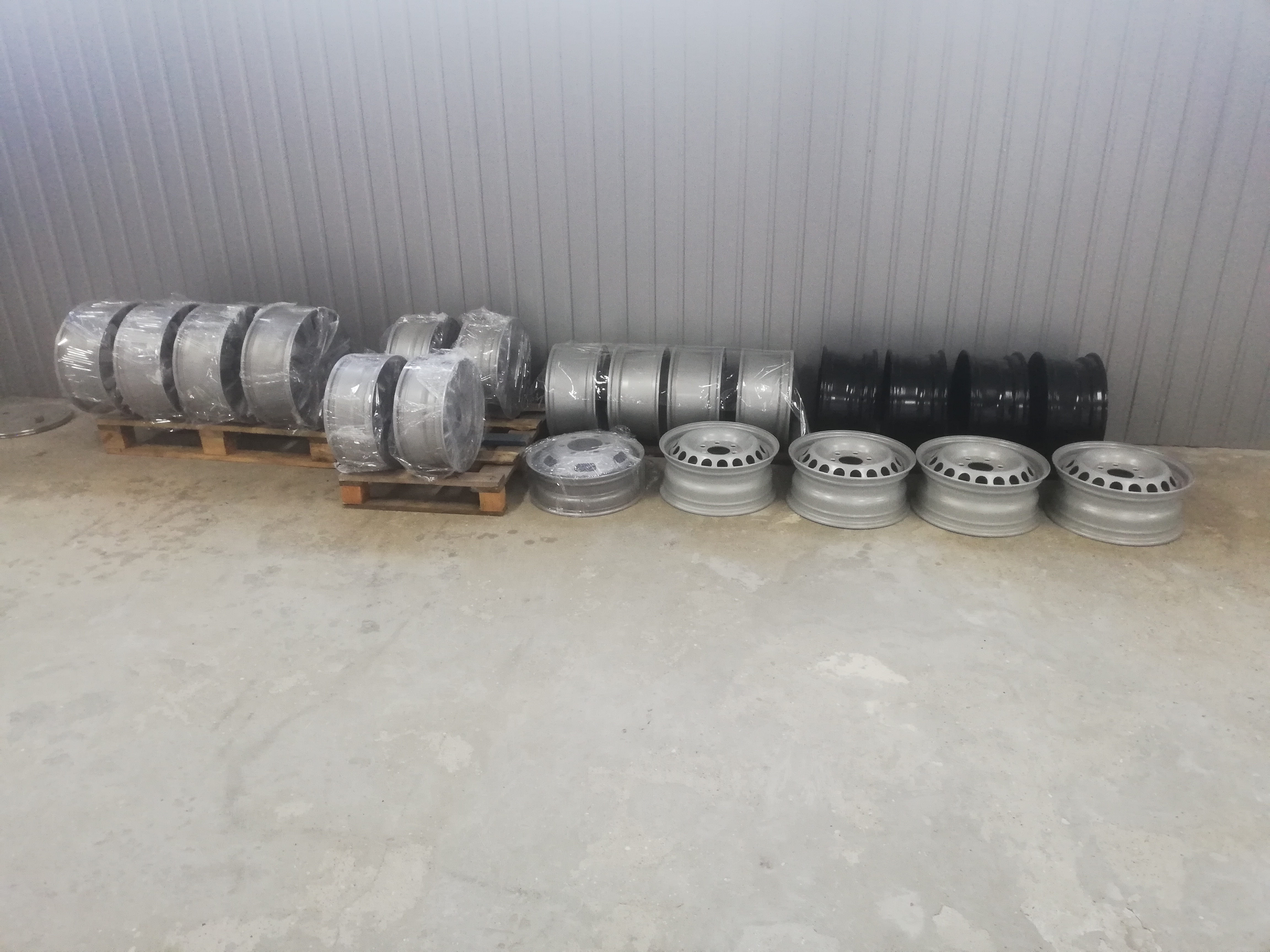 Rzędy felg samochodowych, niektóre owinięte folią, w kolorach srebrnym i czarnym, ułożone na palecie i bezpośrednio na betonowej podłodze w hali z blachy falistej.