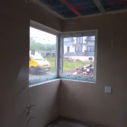 Świeżo otynkowane wnętrze pokoju z widocznymi przewodami elektrycznymi, oknami PCV marki VEKA i żółtym samochodem dostawczym za oknem w deszczowy dzień.